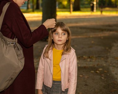 Bir annenin kadın eli ya da bakıcısı dikkatlice küçük bir kızın saçına toka takar. Açık havada sonbahar fotoğrafı. Saç aksesuarları. Dokunaklı kavram