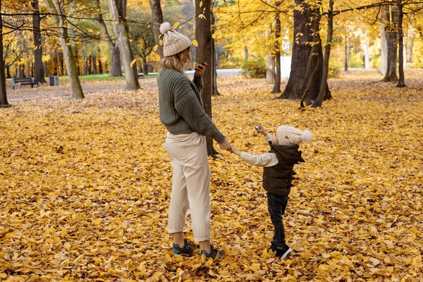 A young active mom is talking on a smartphone, and her child, a little son, needs attention and is naughty in autumn park among yellow foliage. Full length photo