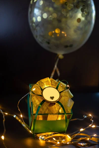 Πράσινο δώρο με χρυσό τόξο με γυαλιστερή διακόσμηση ballwoon με μικρή καρδιά σε σκούρο φόντο με το φως των Χριστουγέννων. Συνέπεια διακοπών. Αντιγραφή χώρου. — Φωτογραφία Αρχείου