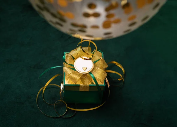 Πράσινο δώρο με χρυσό τόξο με γυαλιστερή διακόσμηση ballwoon με μικρή καρδιά σε σκούρο φόντο με το φως των Χριστουγέννων. Συνέπεια διακοπών. Αντιγραφή χώρου. Φωτογραφία — Φωτογραφία Αρχείου