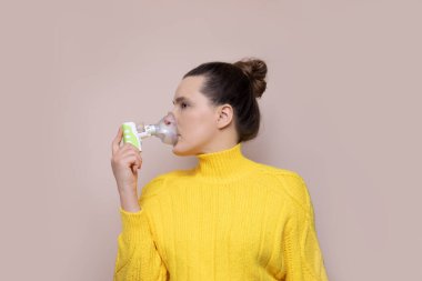 A 40-year-old woman in a yellow sweater on a pink background inhales the upper respiratory tract, lungs with a nebulizer. Looking away. Studio photo. Copy space clipart