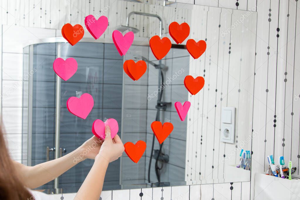 Hands of young girlwoman in the bathroom glue valentine red hearts on mirror in bathroom. Valentines day concept