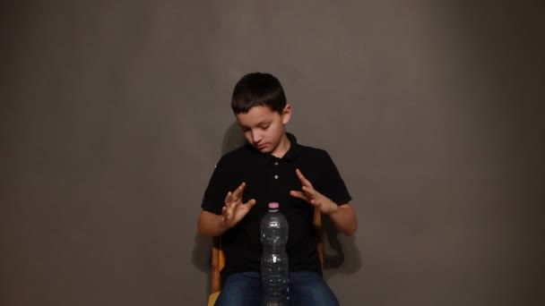 Teenager-Junge zeigt Trick mit Plastikflasche auf grauem Hintergrund. Video mit wegfliegendem Deckel — Stockvideo