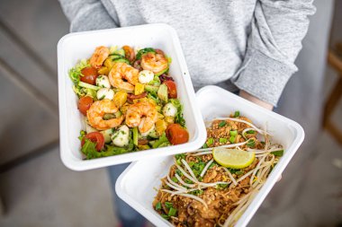 Ofise yemek teslimatı ya da tek kullanımlık plastik tabaklarda piknik için. Teslimatçı elinde karides salatası ve Tayland yemeği tutuyor..