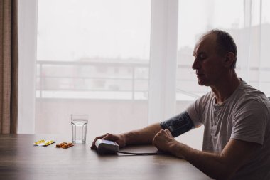 Pencerenin kenarında oturan yaşlı adamın silueti kan basıncını cihazla ölçüyor. Masada bir bardak su ve hap var. Sağlık ve öz bakım konsepti fotoğrafı
