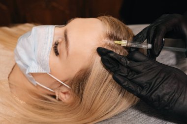 Beautician in gloves gives injection into the head of blonde woman. Mesotherapy, hair loss therapy. clipart