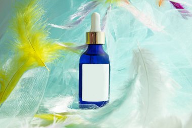 Pipetli mavi cam şişe, mavi tekstil arka planındaki tüyler arasında yüz serumu,