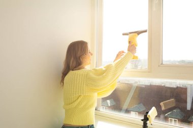 Batan güneş ışınlarındaki genç ev hanımı camları silmek için cihaz kullanıyor. Temiz cam konsepti.