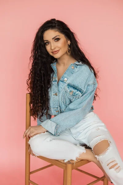 Bella donna in giacca di jeans e jeans strappati siede sulla sedia in studio. concetto di bellezza e cura personale. — Foto Stock