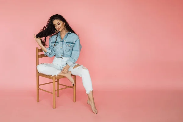 Young beautiful woman with dark curly hair on pink background sits on chair. hair and self care concept.