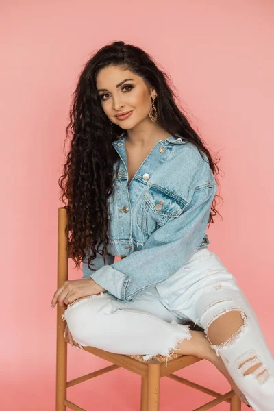 beautiful woman in denim jacket and ripped jeans sits on chair in studio. beauty and personal care concept