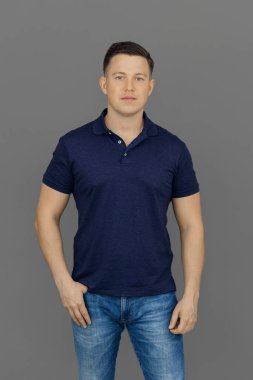 Ciddi genç adam polo gömlek ve kot pantolon içinde gri arka planda duruyor. Kendini ifade etme ve hedeflere ulaşma kavramı.