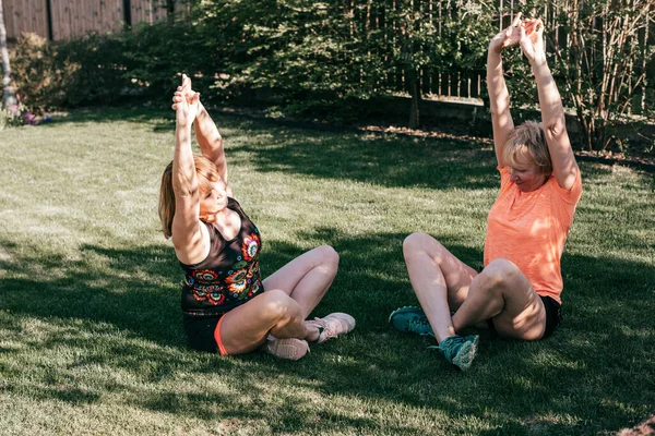 Two senior women on the grass perform stretching during yoga class on summer evening