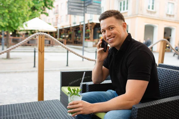 Young businessman man sitting at table of street cafe with cooling mojito cocktail, smiling and looking at camera, using smart phone