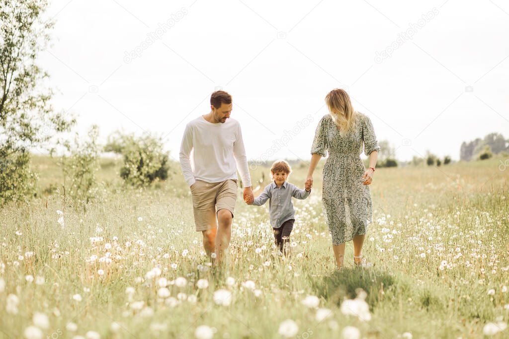 Photo of Cheerful parents having fun with their little son 3 years in summer field while holding hands and walking together.