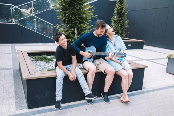 年轻的父亲在城市广场为他的家人在吉他上弹着欢快的歌曲 生活方式 — 图库照片