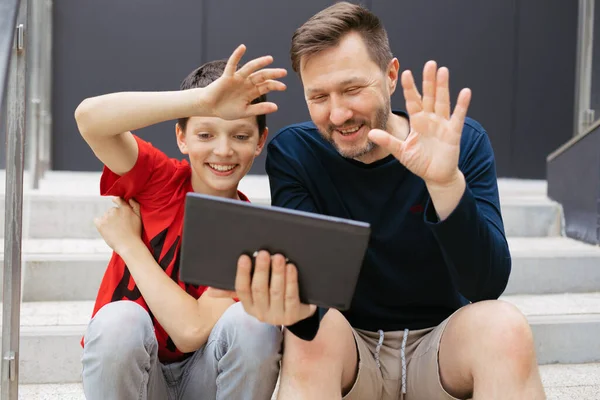 Dad and son make video call using tablet in city on concrete staircase, cheerfully wave their hands saying hello
