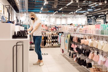 Beyaz tişörtlü ve kotlu sarışın kasaya ödeme yaparken koruyucu maske kullanıyor. Kişisel koruyucu ekipman. COVID-19. Salgın sırasında yapılan eylem. Yaşam Tarzı. Perakende alım satımı. Tüketici malları. 