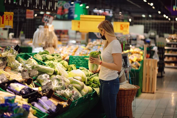 Beautiful young woman in mask chooses vegetables and herbs in supermarket. Proper nutrition. Everyday lifestyle. Natural food. Food products. Personal protective equipment. Social life in epidemic.