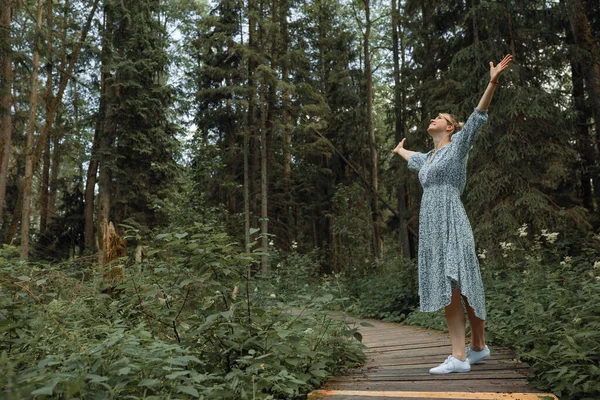 Young beautiful woman breathes fresh air in forest raising her hands up. Concept of outdoor activities. copy space.