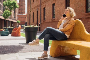  Young woman admires urban architecture while sitting on bench in city and talking on phone. Mobile communication. Tourism and modern technologies. Recreation and entertainment.Modern life.