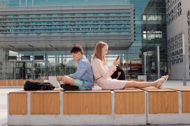 Portrait of cute young students wearing casual, sitting back to back, holding mobile devise in hands, seriously looking at screens, schoolyard near campus