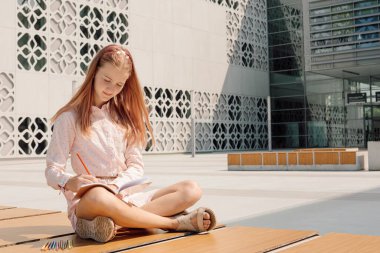 Photo of girl working in notebook with pencil on bench in schoolyard. Informal educational environment
