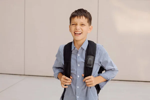 Хлопчик-підліток 11 років школяр дивиться на камеру на сірому фоні з рюкзаком, посміхаючись. Одягнений у сорочку — стокове фото