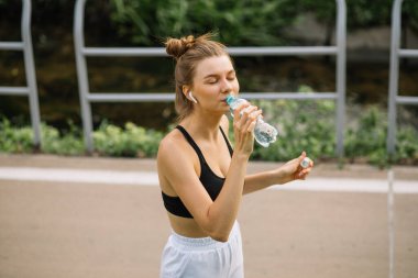 Güzel genç sporcu kadın plastik şişeden su içiyor, spor yaparken susuzluğunu gideriyor.