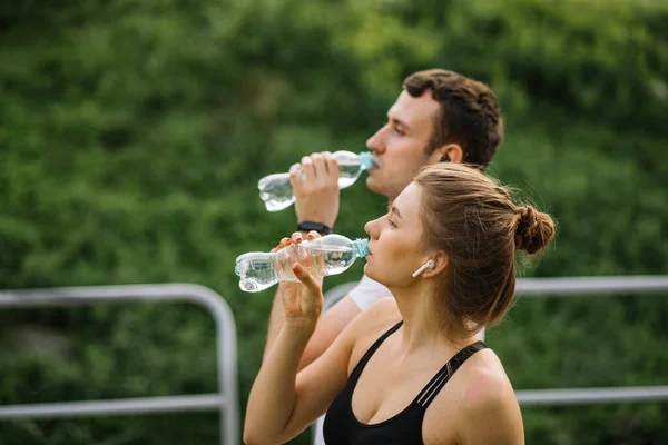 Young happy couple running in city park with plastic botle of water in hands, joint sports, cheerfulness, city sport healthy lifestyle, fitness together, runners, drinking water, thirst —  Fotos de Stock