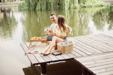 Nehir ya da göl kenarında romantik piknik yapan genç çiftin yan görüntüsü, kadın ve erkek açık havada şarap içiyor, yaz tatilinde eğlenen insanlar, yaşam tarzı.