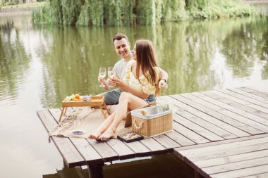 Nehir ya da göl kenarında romantik piknik yapan genç çiftin yan görüntüsü, kadın ve erkek açık havada şarap içiyor, yaz tatilinde eğlenen insanlar, yaşam tarzı fotoğrafı.