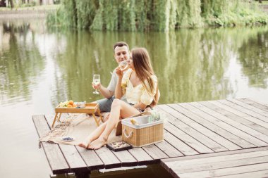 Nehir ya da göl kenarında romantik piknik yapan genç bir çift, kadın ve erkek açık havada şarap içiyor, yaz tatilinde eğlenen insanlar, mutlu bir yaşam tarzı.