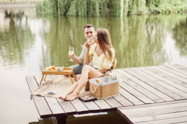 Nehir ya da göl kenarında romantik piknik yapan genç bir çift, kadın ve erkek açık havada şarap içiyor, yaz tatilinde eğlenen insanlar, mutlu bir yaşam tarzı, köy hayatı.