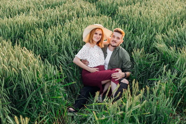 Pregnant young woman sits on her husband\'s lap in field during evening walk. Pregnancy and care. Happiness and tenderness. Care and attention. Love and hope . Healthy lifestyle. Walking in nature.