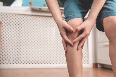 Evde bir çocuğun dizinde zedelenme var. Çocuk elleriyle dizini tutuyor. Bir çocukta diz ağrısı