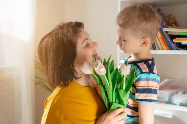 Çocuk, annesine evde bir buket lale çiçeği veriyor. Anneler Günü. Annesi bebeğini seviyor ve ona sarılıyor.