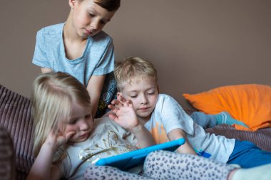 Çocuklar evdeki kanepede otururken tabletle oynarlar. Çocuk bir aletle oyun oynuyor. Çocuklar için tabletteki oyunlar. Çocukların evde boş vakitleri.