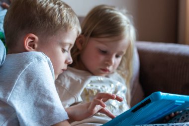 Çocuklar evdeki kanepede otururken tabletle oynarlar. Çocuk bir aletle oyun oynuyor. Çocuklar için tabletteki oyunlar. Çocukların evde boş vakitleri.