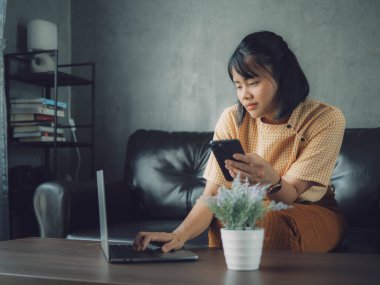 Evde dizüstü bilgisayar ve akıllı telefon kullanan Asyalı kadın. Genç Asyalı iş kadını akıllı telefondan bahsediyor ve labaratuvarla çalışıyor. Ev konsepti üzerine çalışıyor..