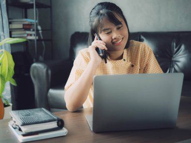 Evde dizüstü bilgisayar ve akıllı telefon kullanan Asyalı kadın. Genç Asyalı iş kadını akıllı telefondan bahsediyor ve labaratuvarla çalışıyor. Ev konsepti üzerine çalışıyor..