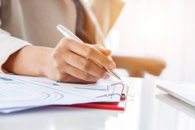 Rapor üzerinde çalışırken not alan finans müdürünün elleri, ofis içinde çalışan kadınlar.