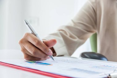 Rapor üzerinde çalışırken not alan finans müdürünün elleri, ofis içinde çalışan kadınlar.