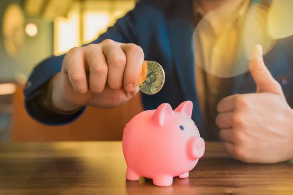 Bir işadamının eli, bir domuz kumbarasının yanında altın bir bitcoin kripto para tutuyor. Bitcoin, kripto para birimi ve tasarruf kavramı.