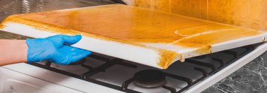 Evdeki lastik eldivenli bir ev kadınının eli kirli bir gaz sobasının kapağını yapışkan bir kaplamayla tutuyor. Mutfağı temizleyin..