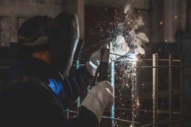 Maskeli bir işçi, bir fabrika veya endüstriyel işletmedeki metal yapılar üzerinde kaynak yapıyor..