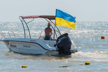 Ukrayna, Demir Liman - 24 Ağustos 2020: Çıplak gövdeli şapkalı genç bir adam, sürat teknesinin sürücüsü sahilde duruyor. Ukrayna ulusal işareti ve sembolü.