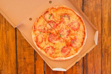 Karton bir paketin içinde peynirli ve salamlı taze yapılmış pizza ahşap bir masanın arka planına karşı zararlı bir fast food..