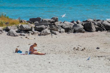 Ukrayna, Demir Liman - 27 Ağustos 2020: Hasır şapkalı, bronzlaşmış yetişkin bir kadın sahildeki dalgakıran taşlarının yanında oturuyor..