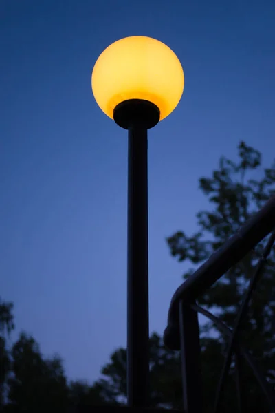 Farola con luz amarilla brillante contra el cielo azul oscuro de la noche. Lámpara exterior en forma de esfera brillante al atardecer. — Foto de Stock